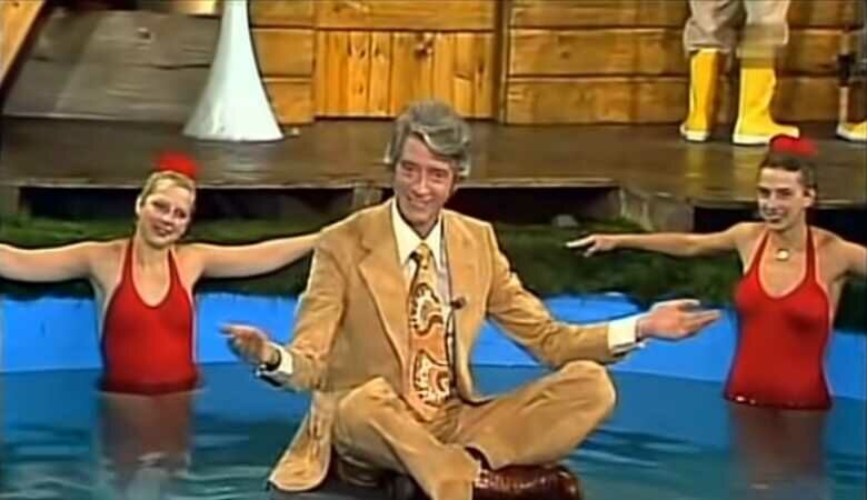 Rudy Carrel in beigen Anzug auf einem Stein in einem Pool sitzend